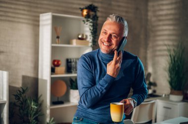 Orta yaşlı işadamı bir ofis masasında oturuyor ve sabah kahvesini içerken akıllı bir telefonla konuşuyor. Sabahları iş görüşmelerini yöneten beyaz bir adam. Boşluğu kopyala.