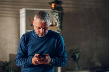 Boğazlı kazaklı orta yaşlı bir adamın portresi. Evde akıllı bir telefona yazı yazıyor. Gri saçlı adam cep telefonu kullanıyor ve iş e-postalarını ya da mesajlarını ailesine tekrar ediyor. Boşluğu kopyala.