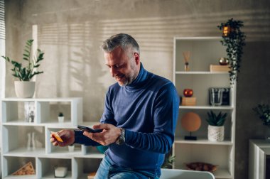 Evde masaya yaslanırken akıllı telefon ve kredi kartı kullanan orta yaşlı yakışıklı bir adam. İnternetten alışveriş yapan, faturaları ödeyen ve online bankacılık yapan gri saçlı bir adam. Mali işler. Boşluğu kopyala.