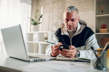 Evde çalışan orta yaşlı bir işadamı akıllı bir telefonla yazı yazıyor. Gri sakallı ve saçları olan beyaz bir adam ev ofisinde çalışırken cep telefonu kullanıyor. Boşluğu kopyala.