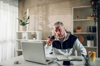 Orta yaşlı iş adamı evde çalışıyor ve video görüşmesi yaparken bir dizüstü bilgisayarı işaret ediyor. Müşterilerle ya da iş arkadaşlarıyla çevrimiçi bir toplantı yapan, arkadaşlarıyla, ailesiyle konuşan bir adam..