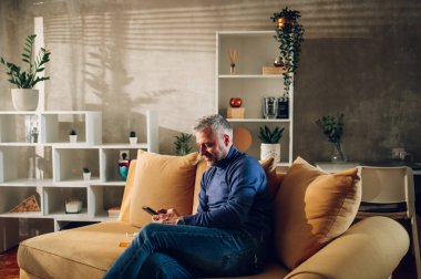 Orta yaşlı, kır saçlı bir adam evde koltukta otururken akıllı telefon kullanıyor. Beyaz adam ev finansmanını yönetiyor, banka hesabını inceliyor ya da cep telefonunda mesaj yazıyor. Boşluğu kopyala.