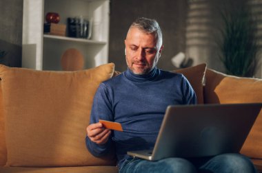 Orta yaşlı, gri saçlı ve sakallı bir adam evde bir kanepede otururken online alışveriş için laptop ve kredi kartı kullanıyor. Faturaları ve kirayı internet bankacılığıyla ödemek, malları satın almak..