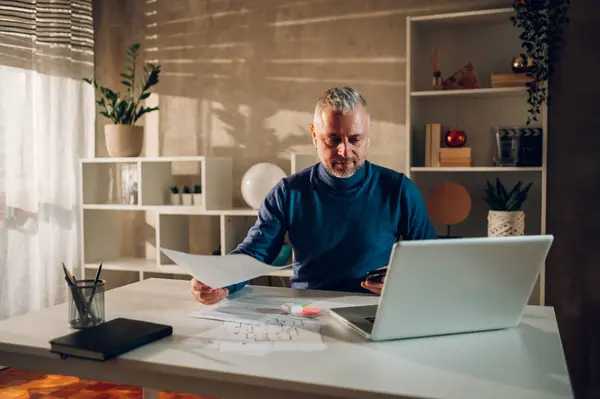 stock image Middle aged businessman working at home while typing on a smartphone and using laptop. Caucasian man working remotely in a home office while sitting at the white table. Copy space.