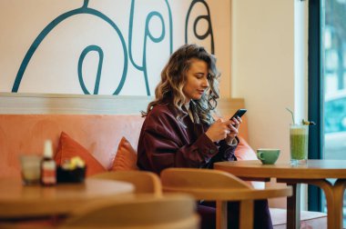 Matcha latte kahve içen ve kafede akıllı telefon kullanan güzel genç bir kadın.