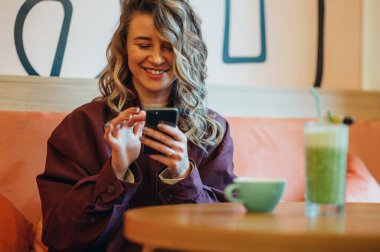 Matcha latte kahve içen ve kafede akıllı telefon kullanan güzel genç bir kadın.