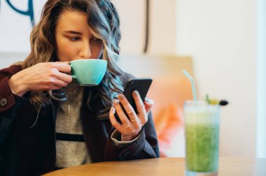 Kahve içen ve kafede akıllı telefon kullanan güzel genç bir kadın.