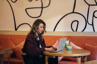 Güzel genç bir kadın kahve içiyor ve bir kafede dizüstü bilgisayarıyla çalışıyor.