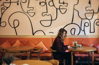 Güzel genç bir kadın kahve içiyor ve bir kafede dizüstü bilgisayarıyla çalışıyor.