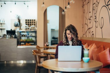 Güzel genç bir kadın kahve içiyor ve bir kafede dizüstü bilgisayarıyla çalışıyor.