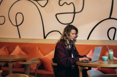 Güzel genç bir kadın kahve içiyor ve bir kafede dizüstü bilgisayarıyla çalışıyor.