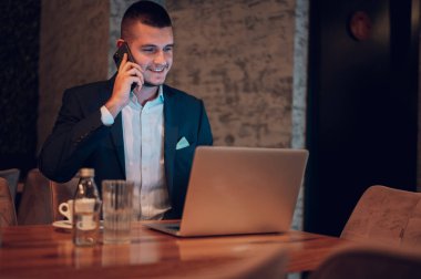 Genç yakışıklı iş adamı akıllı telefon kullanıyor ve bir kafede kahve içerken dizüstü bilgisayarıyla çalışıyor.