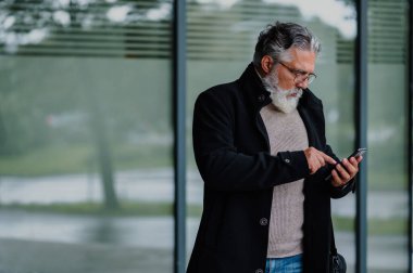 Toplantıya gidip akıllı telefon kullanan, gülümseyen ve kendinden emin bir iş adamının portresi. Ofis binasının dışında şık siyah ceket giymiş yaşlı sakallı adam..
