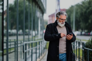 Beyaz sakallı, resmi kıyafetli, caddede yürürken akıllı telefon kullanan gülümseyen kıdemli iş adamı. Olgun bir iş adamı cep telefonuna mesaj ya da e-posta yazıyor. Boşluğu kopyala.