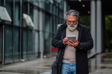 Ofis binasının önünde duran ve bir tablete yazı yazan yaşlı bir işletme yöneticisi. Etrafı binalarla çevrili, teknoloji ve internet kullanan kıdemli bir işadamı..