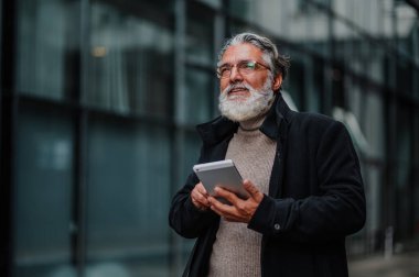 Üst düzey yönetici iş merkezinin önünde tablet kullanıyor. Ofis binasının önünde duran mutlu yaşlı işadamı iş toplantısından önce teknoloji ve internet kullanıyor..