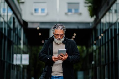 Bir şirket binasının önünde dikilirken dijital tablet kullanan başarılı bir kıdemli işadamının portresi. İş toplantısından sonra ofis dışında yaşlı yönetici..