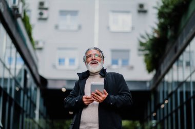 Beyaz sakallı ve resmi kıyafetli gülümseyen kıdemli işadamı ofis binasının yanında dururken tablet kullanıyor. Bir toplantıya hazırlanırken teknolojiyi ve interneti kullanan yaşlı bir adam.