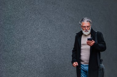 Üst düzey gri saçlı işadamı ofisin dışında duruyor ve akıllı telefon kullanırken gülümsüyor. Yaşlı patron e-posta okuyor ya da cep telefonunda mesaj yazıyor. Arka plandaki gri duvar. Boşluğu kopyala.