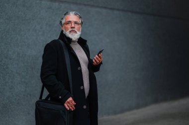 Üst düzey yönetici dışarıda dururken akıllı telefonu kullanıyor. Mutlu yaşlı işadamı gri duvarın önünde duruyor ve cep telefonundan bir mesaj ya da e-posta okuyor. Boşluğu kopyala.
