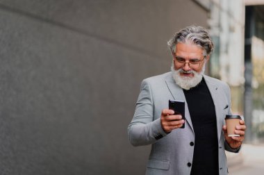 Beyaz sakallı, gri takım elbiseli, arka planda gri bir duvarla sokakta yürürken akıllı telefon kullanan kıdemli, güler yüzlü bir işadamı. Cep telefonundan e-posta okuyan yaşlı bir adam.
