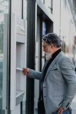 Üst düzey yönetici kredi kartı ya da banka kartı kullanıp bankadan para çekiyor. ATM makinesiyle gri takım elbiseli mutlu bir iş adamı. Modern bankacılık. Boşluğu kopyala.