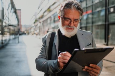 Üst düzey yönetici bir panoya not alıyor ve iş merkezindeki toplantıya gidiyor. Ofis binasının önünde duran ve iş fikirleri yazan mutlu yaşlı işadamı. Boşluğu kopyala.
