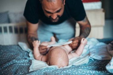 Dövmeli sakallı baba evde yatağında oğluyla oynarken bez takıyor.