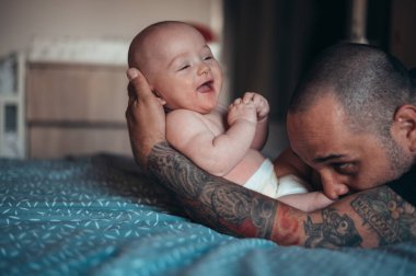 Dövmeli sakallı baba evde yatağında oğluyla oynarken bez takıyor.