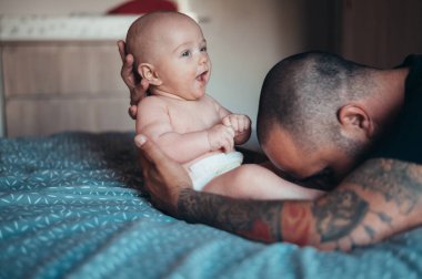 Dövmeli sakallı baba evde yatağında oğluyla oynarken bez takıyor.