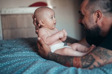 Dövmeli sakallı baba evde yatağında oğluyla oynarken bez takıyor.