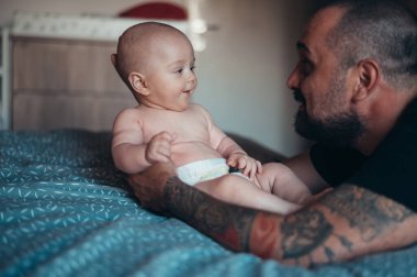 Dövmeli sakallı baba evde yatağında oğluyla oynarken bez takıyor.