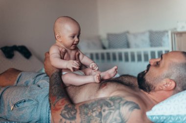 Dövmeli, sakallı, ispanyol bir baba evdeyken oğluyla bebek bezi ile oynuyor.