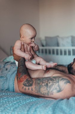 Dövmeli sakallı baba evde yatağında oğluyla oynarken bez takıyor.