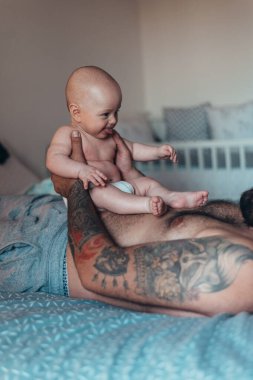 Dövmeli sakallı baba evde yatağında oğluyla oynarken bez takıyor.