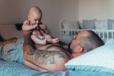 Dövmeli sakallı baba evde yatağında oğluyla oynarken bez takıyor.