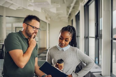 Resmi giyinmiş iki çok ırklı iş arkadaşı mola sırasında kahve içip gazete okuyorlar. Neşeli Afrika kökenli Amerikalı kadın menajer, beyaz çalışanıyla iletişim kuruyor..