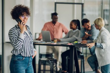 Ofiste duran ve arka planda çalışan iş arkadaşlarıyla akıllı telefondan konuşan pozitif kadın yöneticiler. Ofiste afro saçlı, gülümseyen İspanyol iş kadını..