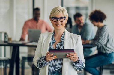 Ofiste gözlüklü gülümseyen bir iş kadını ve arka planda iş arkadaşlarıyla buluşurken tablet kullanıyor. Başarılı kadın profesyonel. Sarışın kadın kameraya bakıyor..