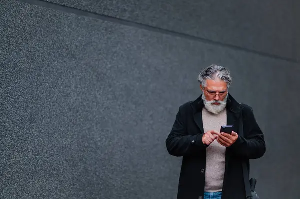 Yetişkin bir iş adamının portresi dışarıda duruyor ve bir toplantıya giderken akıllı bir telefonla daktilo yazıyor. Üstün sakallı, şık, siyah paltolu bir adam gri bir duvarın önünde dikiliyor. Boşluğu kopyala.