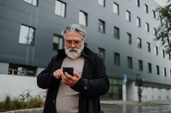 Ofis binasının önünde duran ve cep telefonu kullanan olgun bir yöneticinin portresi. Sokaklarda binalarla çevrili kıdemli bir işadamı duruyor ve akıllı bir telefonla daktilo yazıyor.