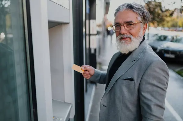 Kredi kartı tutan ve ATM 'nin önünde duran kıdemli yakışıklı işadamı ya da girişimci. Başarılı yaşlı bir adam banka hesabından para çekiyor. Modern bankacılık. Boşluğu kopyala.