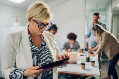 Meslektaşları arka planda bir toplantı düzenlerken, kendine güvenen bir ifadeyle poz veren güzel genç bayan takım lideri. Konferans odasında iş arkadaşları olan olumlu bir kadın. Dijital tablet kullanan iş kadını