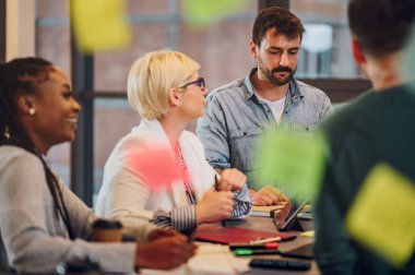 Modern, endüstriyel tasarım ofisinin içindeki bir toplantıda çok ırklı bir iş ekibi. Takım lideri projeyi çeşitli meslektaşlarına sundu. Üzerinde çıkartmalar olan cam bir duvarın içinden bak..