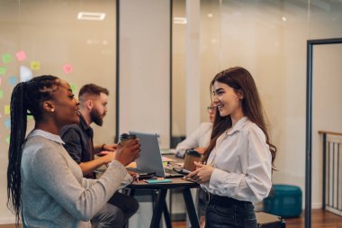 Bir grup çok ırklı genç meslektaşın toplantı odasında şirket projeleri hakkında konuşuyorlar. Takım çalışması konsepti. Çeşitli iş arkadaşları birlikte çalışıyor. İki bayan meslektaşına odaklan..
