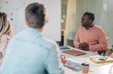 Çok kültürlü profesyonel proje yöneticileri grubu toplantı salonunda müzakere ediyor. Toplantıda iş arkadaşını dinleyen Afro-Amerikalı bir adama odaklan. Boşluğu kopyala.