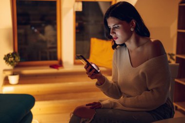 Güzel bir kadın hafta sonları evde dinlenirken akıllı telefon kullanıyor ve cihaz ekranı tarafından aydınlatılıyor. Gecenin bir yarısı akıllı telefondan medyanın içeriğini izlemek.