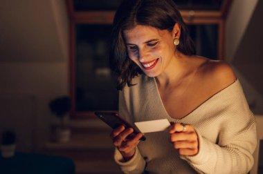 Akıllı telefon ve kredi kartı kullanan beyaz güzel bir kadının portresi gece evde dinlenirken ve cihaz ekranı tarafından aydınlatılırken. Evden online alışveriş..