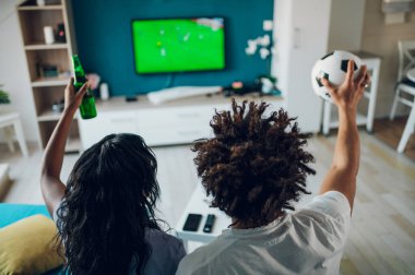 Afrikalı Amerikalı spor fanatiği çift TV 'de önemli futbol maçlarını izlerken evde bira içip tezahürat yapıyorlar. Futbol topu tutan adam. Favori bir takım için tezahürat. Arka görünüm.