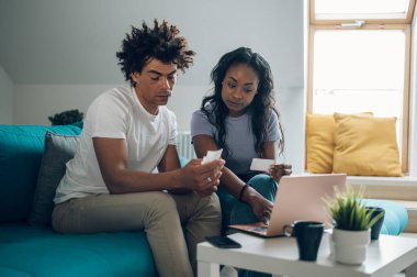 Afrikalı Amerikalı çift hesaplarını evde yapıyorlar ve online ödeme için bir laptop ve kredi kartı kullanıyorlar. İnternetten alışveriş ve alım. Faturalar ve aile bütçesi.